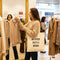 Woman in a clothing store holding a beige tote bag with text, surrounded by racks of clothes.