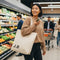 Woman holding an avocado and a reusable tote bag with 'J.B.' in a grocery store.