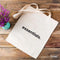 Beige tote bag with 'essentials.' text on a wooden surface with a small plant.