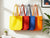 Four colorful tote bags (yellow, red, blue, orange) hanging on a rack against a white wall.